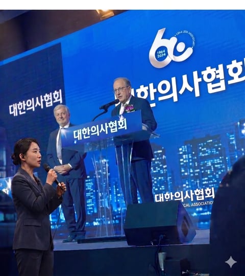 Man in business suit speaking at podium on stage with blue backdrop and Korean text, woman in foreground holding microphone
